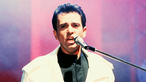 Mężczyzna w białej kurtce śpiewa do mikrofonu na tle kolorowych świateł.