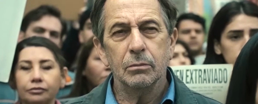 Mężczyzna z krótkimi włosami i zmarszczonym czołem stoi w tłumie protestujących ludzi.