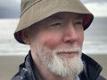 Mężczyzna w kapeluszu na plaży, z siwą brodą i uśmiechem, piękne morze w tle.