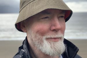 Mężczyzna w kapeluszu na plaży, z siwą brodą i uśmiechem, piękne morze w tle.