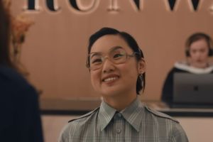 Na zdjęciu uśmiechnięta młoda kobieta w okularach, w plaidzie, w recepcji. W tle druga osoba przy komputerze.