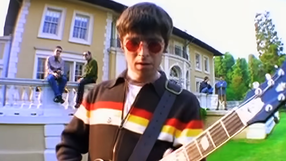 Mężczyzna grający na gitarze przed willą, z trzema osobami siedzącymi na tarasie.