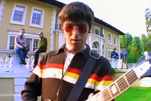 Mężczyzna grający na gitarze przed willą, z trzema osobami siedzącymi na tarasie.