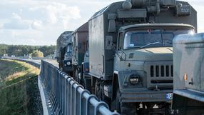 Na zdjęciu widać kolumnę wojskowych ciężarówek na drodze, otoczoną barierkami.