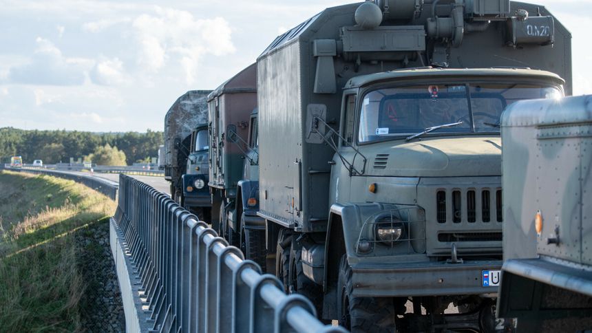 Na zdjęciu widać kolumnę wojskowych ciężarówek na drodze, otoczoną barierkami.