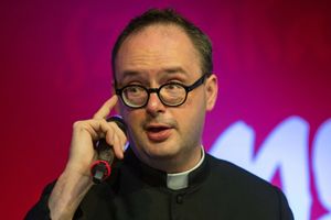 Mężczyzna w okularach, w czarnym stroju, mówi do mikrofonu na tle kolorowej ściany.