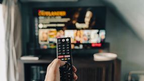 Zdjęcie przedstawia rękę trzymającą pilot, skierowaną w stronę włączonego telewizora.