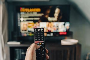 Zdjęcie przedstawia rękę trzymającą pilot, skierowaną w stronę włączonego telewizora.