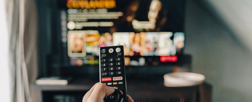 Zdjęcie przedstawia rękę trzymającą pilot, skierowaną w stronę włączonego telewizora.