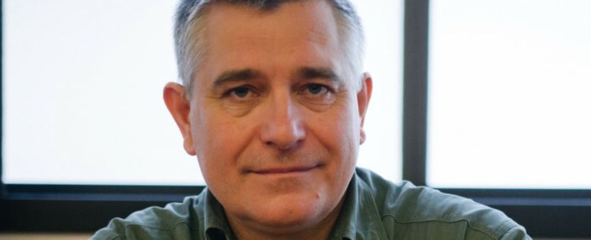 Mężczyzna w zielonej koszuli, siedzi przy biurku, z oknem w tle, spokojny wyraz twarzy.