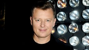 Mężczyzna w czarnej koszulce, uśmiechający się, na tle jasnych świateł.