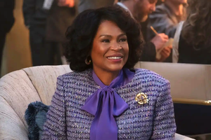 Na zdjęciu uśmiechająca się kobieta w fioletowej bluzce i eleganckim żakiecie siedzi na kanapie.