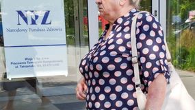 Na zdjęciu widać starszą kobietę w granatowej bluzce w białe i różowe kropki, stojącą przed budynkiem.