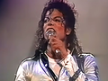 Michael Jackson trzyma mikrofon i śpiewa na scenie w błyszczącej kurtce.