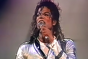 Michael Jackson trzyma mikrofon i śpiewa na scenie w błyszczącej kurtce.