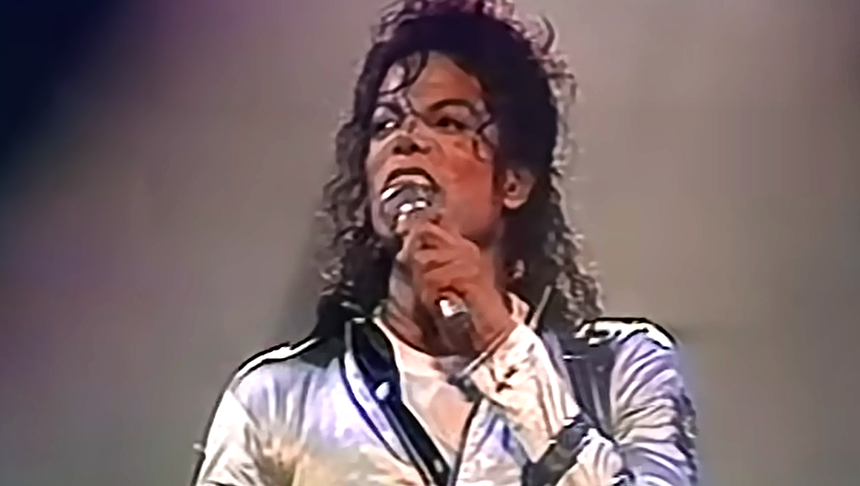 Michael Jackson trzyma mikrofon i śpiewa na scenie w błyszczącej kurtce.