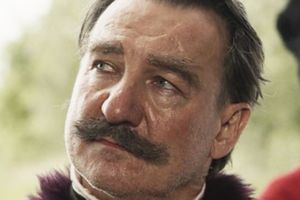 Mężczyzna z wąsami w eleganckim stroju, z poważnym wyrazem twarzy, w plenerze.