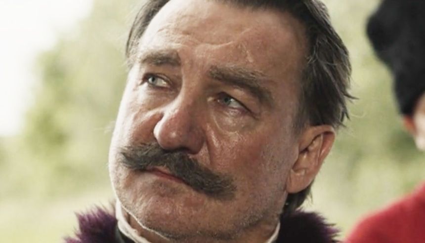 Mężczyzna z wąsami w eleganckim stroju, z poważnym wyrazem twarzy, w plenerze.