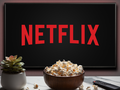 Telewizor wyświetlający logo Netflix, obok miska popcornu i pilot. Roślina doniczkowa w tle.