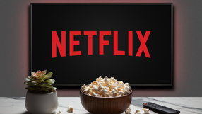 Telewizor wyświetlający logo Netflix, obok miska popcornu i pilot. Roślina doniczkowa w tle.