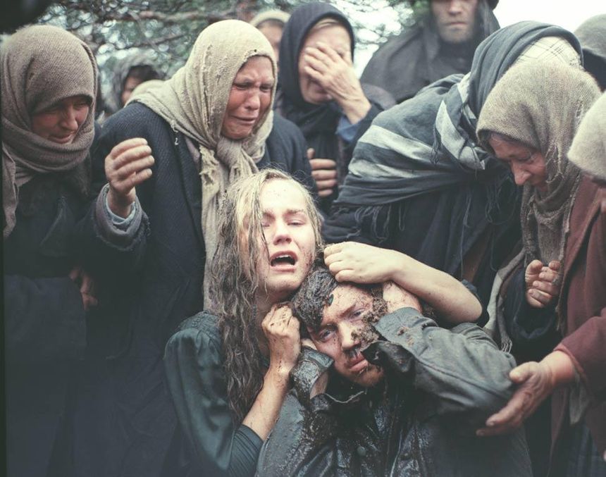 Grupa osób w żalu, kobieta trzyma zmarłego mężczyznę, scenka pełna emocji i smutku.