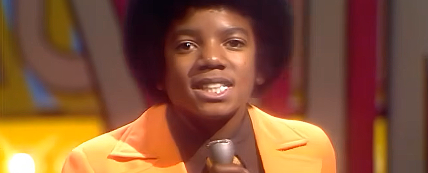 Młody mężczyzna w pomarańczowym garniturze trzyma mikrofon. Śpiewa na scenie.