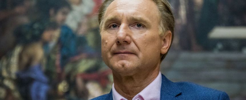 Mężczyzna w jasnej koszuli i granatowym żakiecie, poważnie spogląda w stronę kamery.