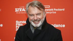 Na zdjęciu uśmiechnięty mężczyzna w ciemnym płaszczu na tle czerwonej ściany z napisami.