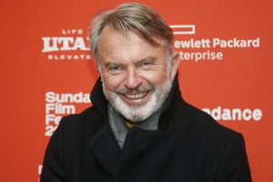 Na zdjęciu uśmiechnięty mężczyzna w ciemnym płaszczu na tle czerwonej ściany z napisami.