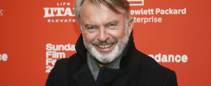 Na zdjęciu uśmiechnięty mężczyzna w ciemnym płaszczu na tle czerwonej ściany z napisami.