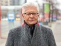 Mężczyzna w szarej kurtce stoi na ulicy, w tle przystanek i zamazane światła pojazdów.