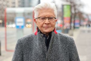 Mężczyzna w szarej kurtce stoi na ulicy, w tle przystanek i zamazane światła pojazdów.