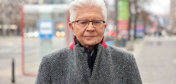 Mężczyzna w szarej kurtce stoi na ulicy, w tle przystanek i zamazane światła pojazdów.