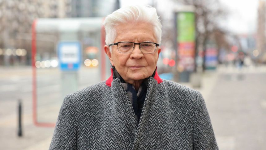 Mężczyzna w szarej kurtce stoi na ulicy, w tle przystanek i zamazane światła pojazdów.