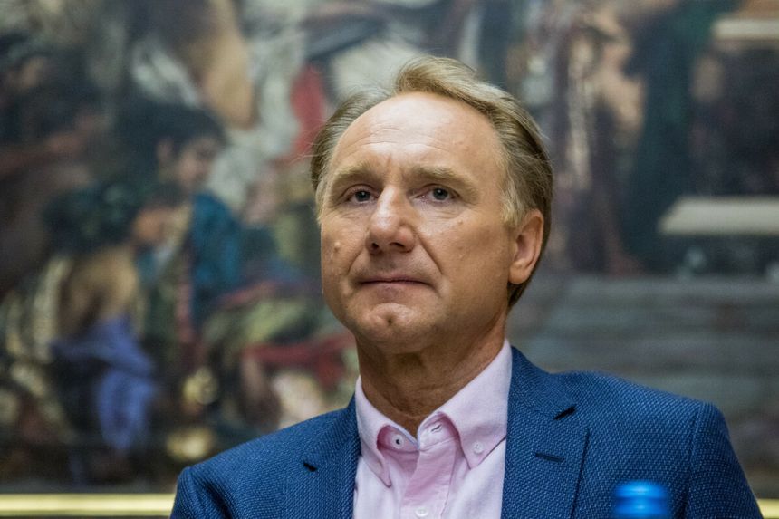 Mężczyzna w niebieskim garniturze, siedzi z poważnym wyrazem twarzy.