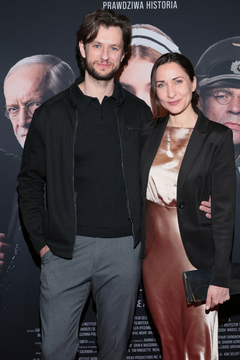 Para ludzi stoi obok siebie na czerwonym dywanie, ubrana elegancko, w tle plakat filmowy.