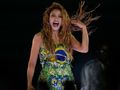 Na zdjęciu znajduje się Shakira, śpiewająca z uśmiechem i falującymi włosami, w kolorowej sukience.