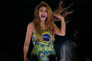 Na zdjęciu znajduje się Shakira, śpiewająca z uśmiechem i falującymi włosami, w kolorowej sukience.