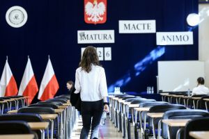 Kobieta w białej bluzce idzie do sali maturalnej, w tle polskie flagi i napisy 