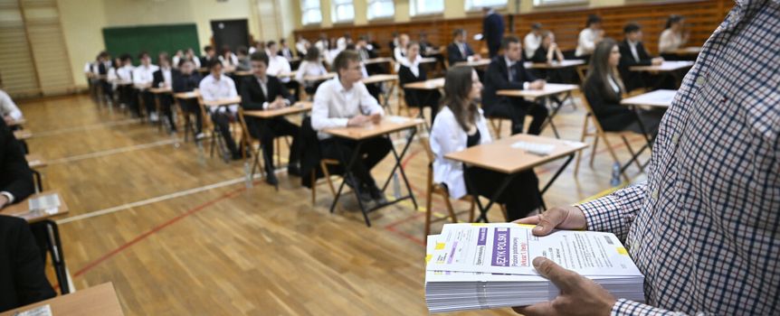 Uczniowie podczas egzaminu w sali gimnastycznej, nauczyciel rozdaje materiały egzaminacyjne.
