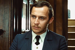 Mężczyzna w eleganckim garniturze, z poważnym wyrazem twarzy, w drewnianym pomieszczeniu.