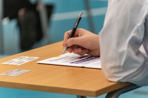 Osoba pisząca długopisem na dokumencie umieszczonym na stole. W tle lekko rozmyte postacie.