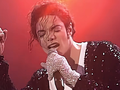 Michael Jackson w czarnym, błyszczącym ubraniu. Śpiewa, trzymając mikrofon i wskazując palcem w górę.