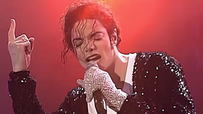Michael Jackson w czarnym, błyszczącym ubraniu. Śpiewa, trzymając mikrofon i wskazując palcem w górę.