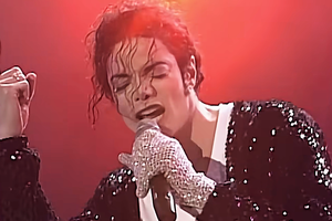 Michael Jackson w czarnym, błyszczącym ubraniu. Śpiewa, trzymając mikrofon i wskazując palcem w górę.