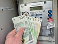 Mężczyzna trzyma banknoty 100-złotowe przed licznikiem energii elektrycznej.
