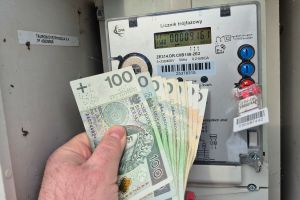 Mężczyzna trzyma banknoty 100-złotowe przed licznikiem energii elektrycznej.