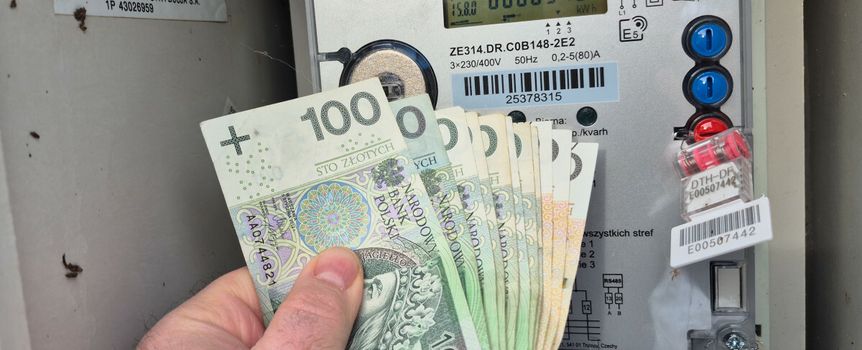 Mężczyzna trzyma banknoty 100-złotowe przed licznikiem energii elektrycznej.