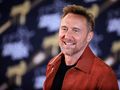 David Guetta z zarostem w brązowej kurtce uśmiecha się przed czarnym tłem z jasnymi napisami.
