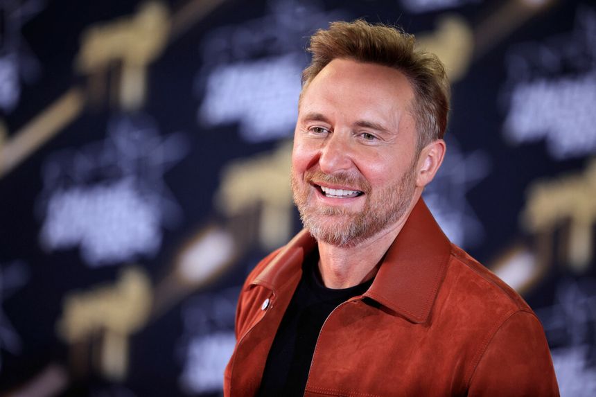 David Guetta z zarostem w brązowej kurtce uśmiecha się przed czarnym tłem z jasnymi napisami.